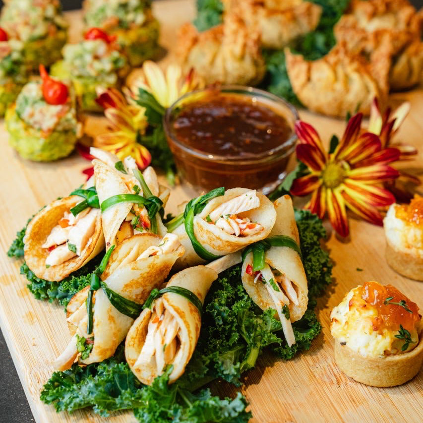 A wooden serving board filled with assorted appetizers, including rolled spring rolls on kale, small stuffed pastries, and a small bowl of dipping sauce, styled with decorative flowers on a dark surface.